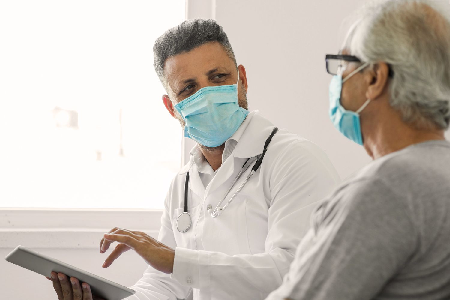 A physician, an Indian man, in a white coat and mask, using a tablet to talk to his patient, an Asian elderly man.