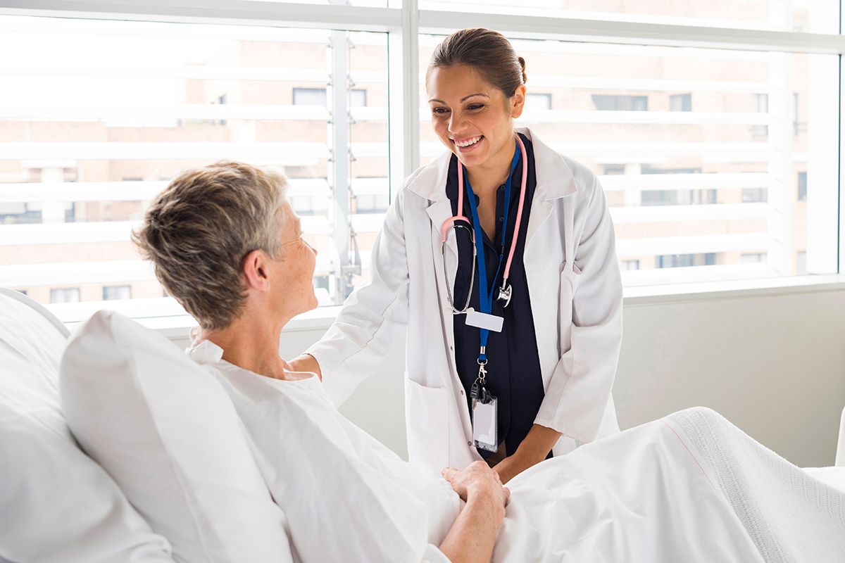 An Internal Medicine physician in a white coat, checking on her patient, a white woman, at a hospital bed and wearing a gown.