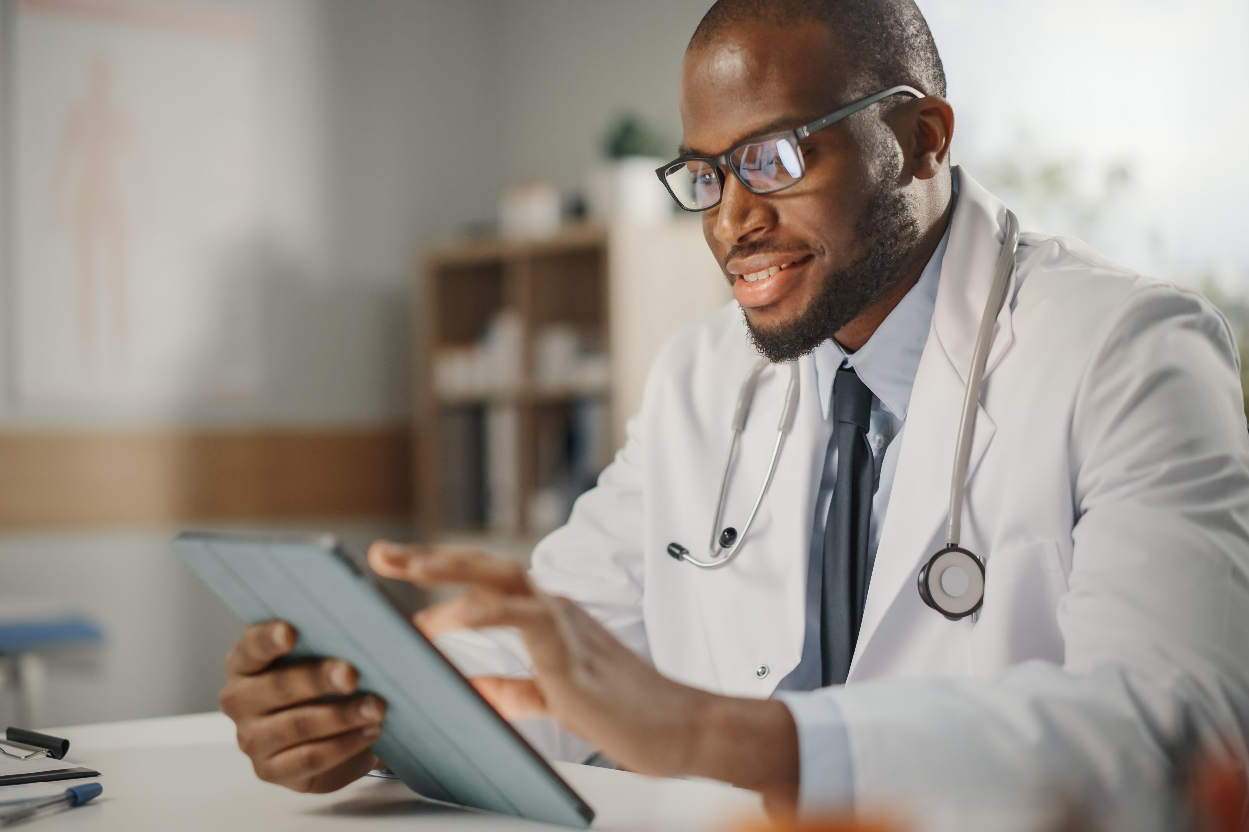 An Oncologist, a Black man, wearing a white coat and stethoscope, smiling as he reviews patient medical records on a tablet.