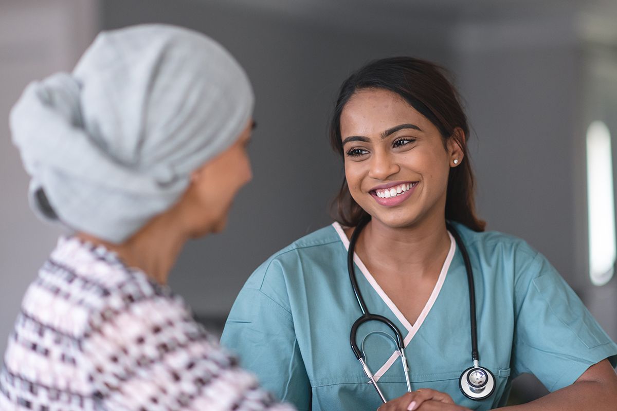 An Oncologist, a Black woman, in blue scrubs and stethoscope, smiling with her patient, an Asian woman in a headwrap.
